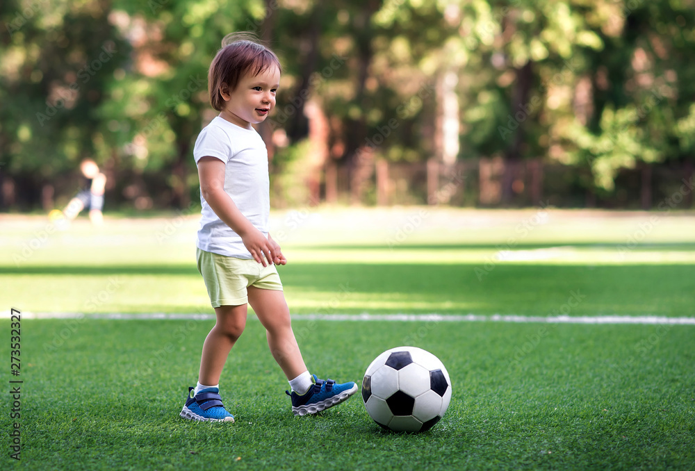 Little football player: toddler boy in sports uniform playing footbal at soccer field in summer day outdoors. Child ready to kick the ball. Active childhood concept Little football player: toddler boy in sports uniform playing footbal at soccer field in summer day outdoors. Child ready to kick the ball. Active childhood concept