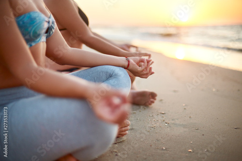 Two women doing group meditation on the beach on sunrise. Female friends doing yoga padmasana or lotus asana sitting on the sand. Girls in swimsuit and leggings relaxing on sunset near sea.