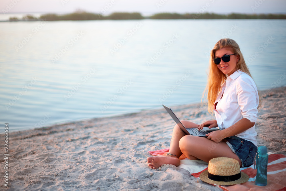 Authentic woman working with laptop on sea shore. Freelancer girl telecommuting with team on project on tropical island. Female using internet from computer while sitting on beach towel on sunset.