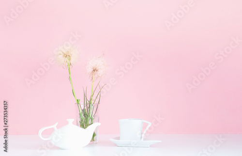 White porcelain teapot and cup on pink background with white dandelions. Concept for festive background.