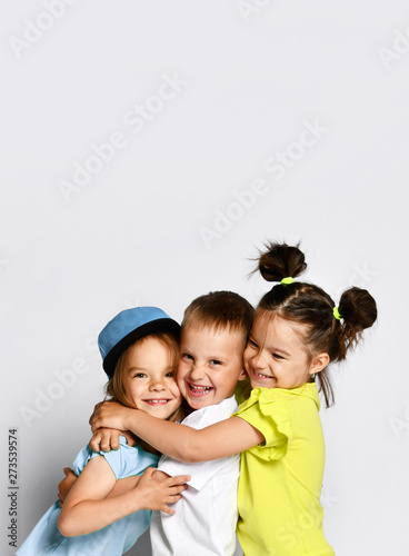 Studio portrait of children on a light background: full body shot of three children in bright clothes, two girls and one boy. Triplets, brother and sisters. hugging on camera. Family ties, friendship