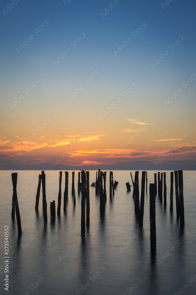 Rusty piles stick out of the water against the background of the sea colorful sunset. Long exposure. Vertical shot.