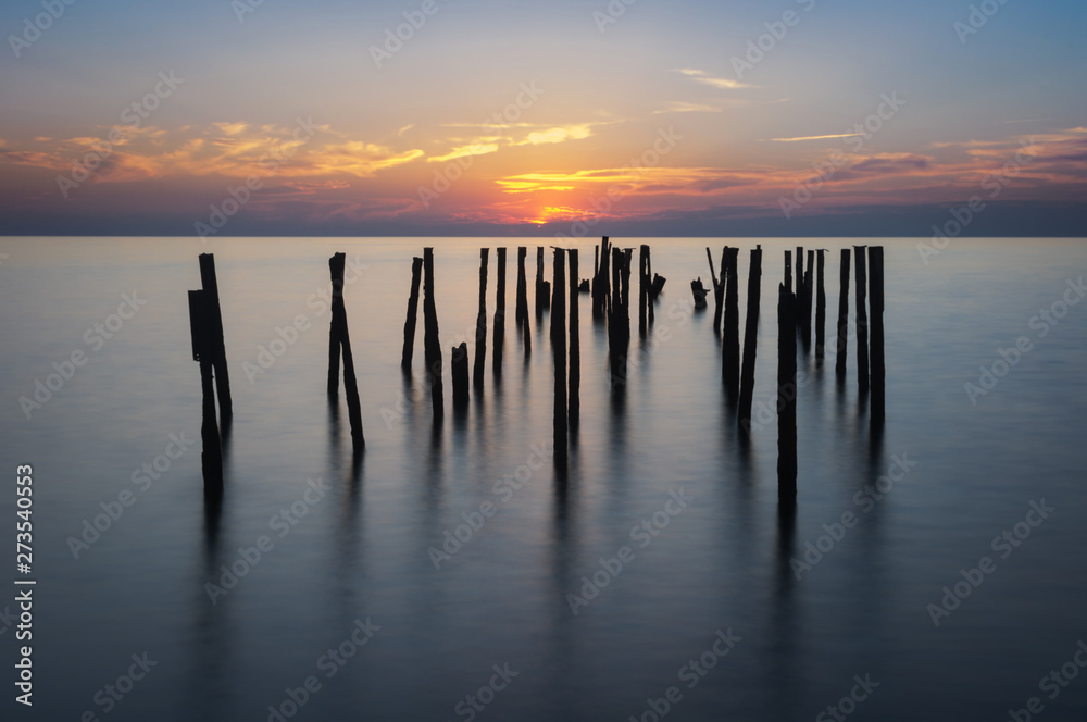 Rusty piles stick out of the water against the background of the sea colorful sunset. Long exposure. Horizontal shot.