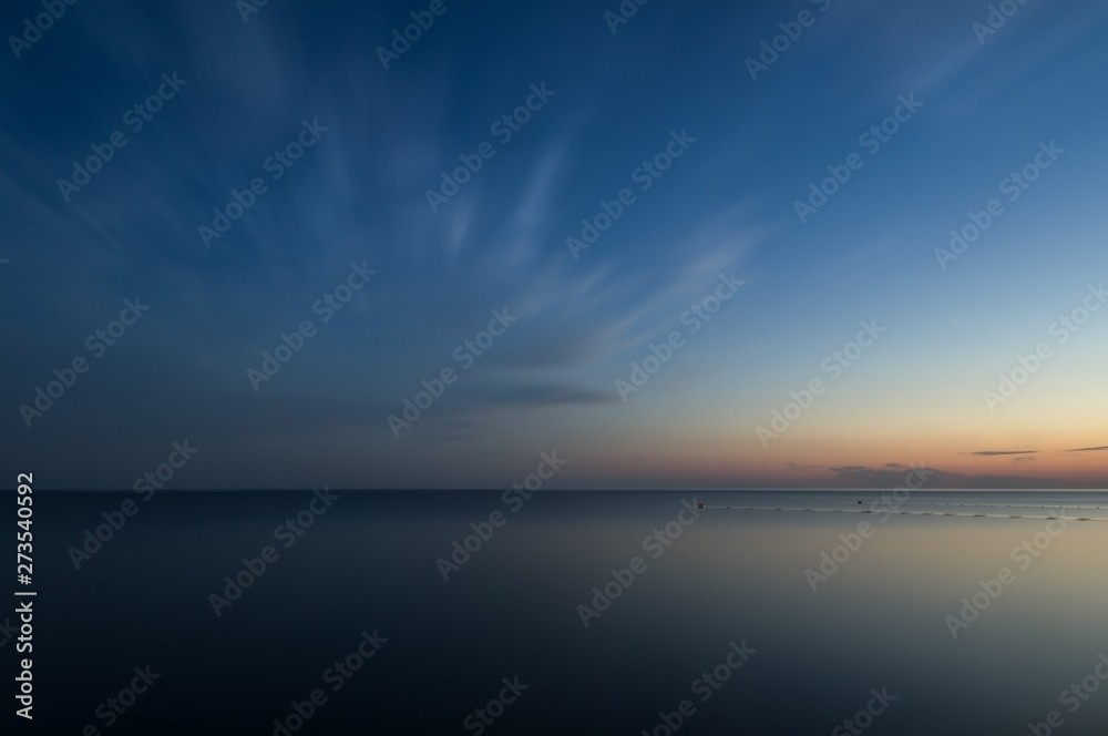 Long exposure. Evening sea under the clouds. The surface of the water turned into a flat surface. Blue sky on water reflections of the sunset. The blurred fence in the water. - obrazy, fototapety, plakaty 