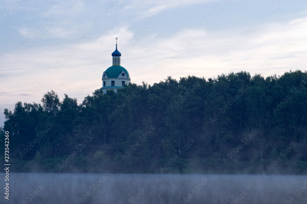 Church among trees on the shore of the morning lake. Fog