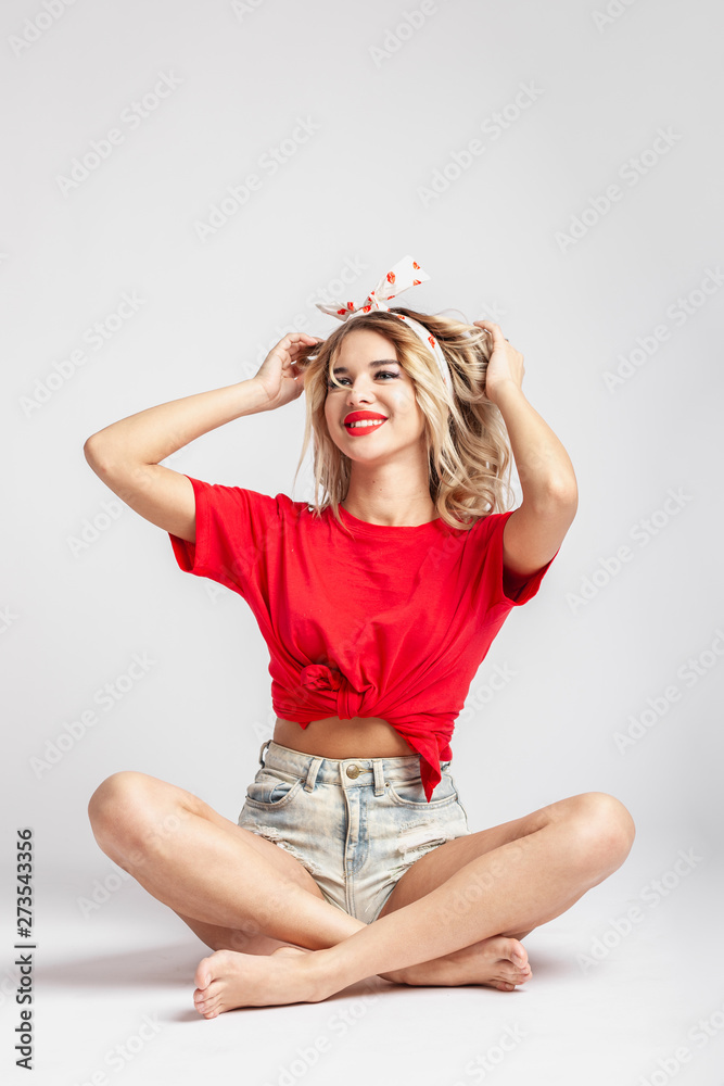 Young blond girl with a ribbon on her head dressed in short denim shorts  and a red t-shirt poses sitting on the floor against a wall in the studio  Stock Photo