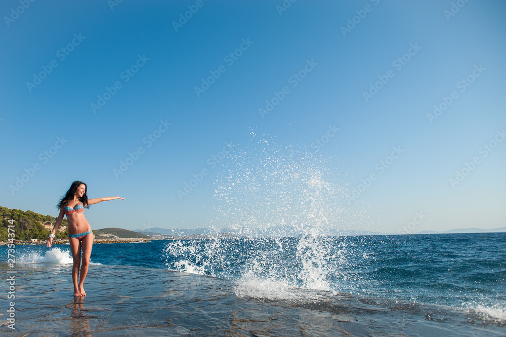 A girl in a swimsuit, splashing from the waves, Brunette girl rejoices splashing from the waves. Young happy woman jumping with raised arms, waving a blue bikini swimsuit in a blue swimsuit and a