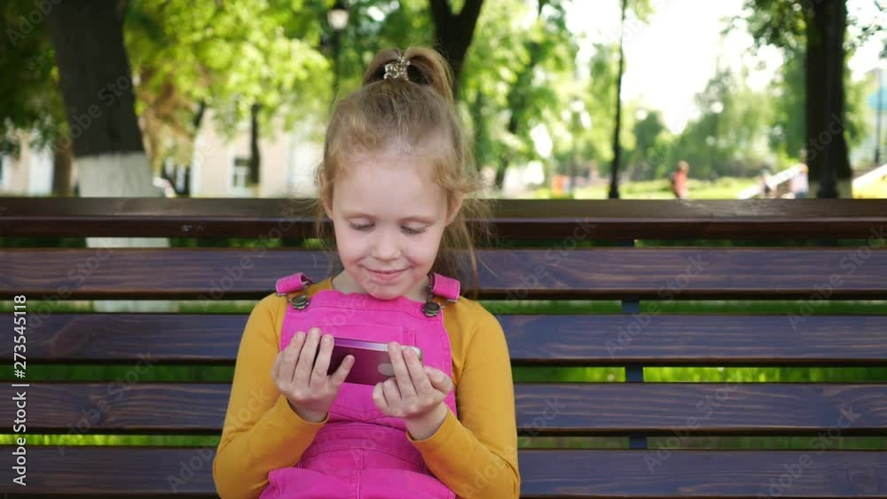 A smiling girl a preschool age , uses the phone outdoors in the park. Sunny summer day. A smiling girl a preschool age , uses the phone outdoors in the park. Sunny summer day.