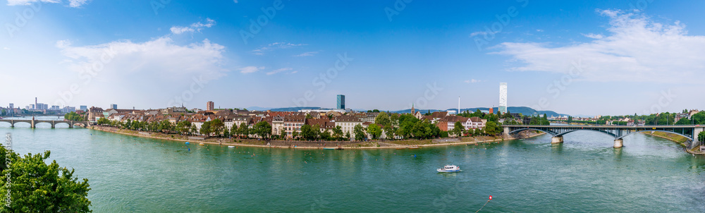 Outdoor sunny panoramic scenery above Rhine river of promenade and bridge along riverside, Basel's cityscape, and people float on river, from terrace behind Cathedral in Summer in Basel, Switzerland.