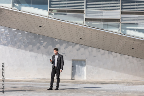 Businessman using mobile phone app texting outside of office in urban modern city building background. Young caucasian man holding smartphone for business work.