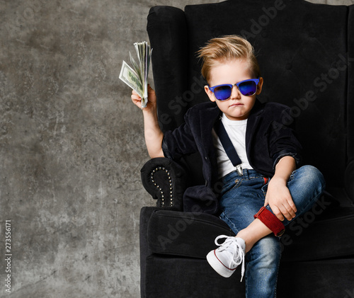Happy and shoutting rich kid boy millionaire sits with a bundle of money dollars cash in big luxury armchair while bills falling photo