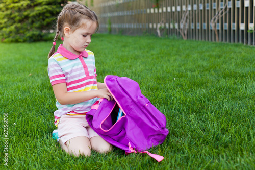 First day of school. child girl schoolgirl elementary school student sitting on the grass near the school and puts books in his backpack. Concept back to school. outdoor activities