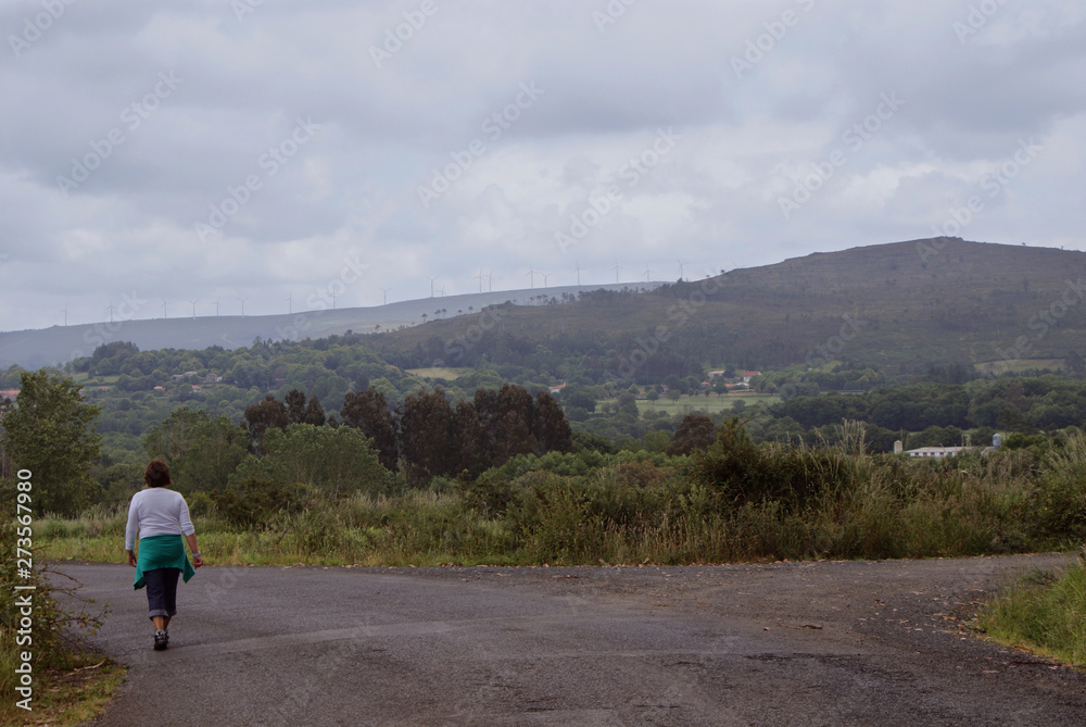 Mujer de mediana edad caminando por ruta de senderismo en día nublado de primavera por carretera rodeada de bosque verde, vegetación, colinas y aerogeneradores al fondo en aldea de Melide, Galicia.