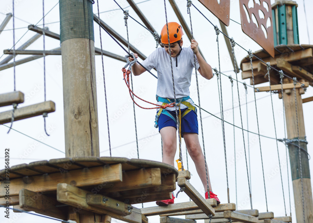 Boy in forest adventure park. Kid in orange helmet and white t shirt climbs on high rope trail. Climbing outdoor, amusement center for children. Young boy plays outdoors	