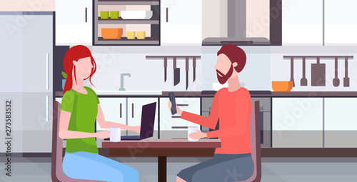 couple sitting at table woman using laptop man holding smartphone digital gadget addiction concept modern kitchen interior flat portrait horizontal