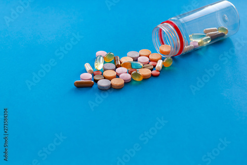 Multi-colored vitamins in capsules and tablets poured from a bottle, on a blue background.