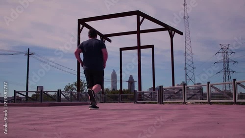 A man jogging into the frame along the Cuyahoga County towpath train near the Cuyahoga River in downtown Cleveland Ohio. Man running in slow motion with a cityscape skyline in the background.