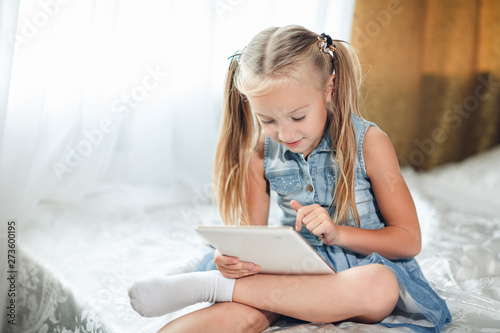 Cute little child girl blond in denim sundress lies in bed uses digital tablet. child playing on tablet pc.