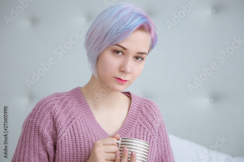 A beautiful young woman with blue hair is sitting on a bed in a bedroom dressed in a lilac, purple sweater, holding a mug with hot coffee or tea. photo