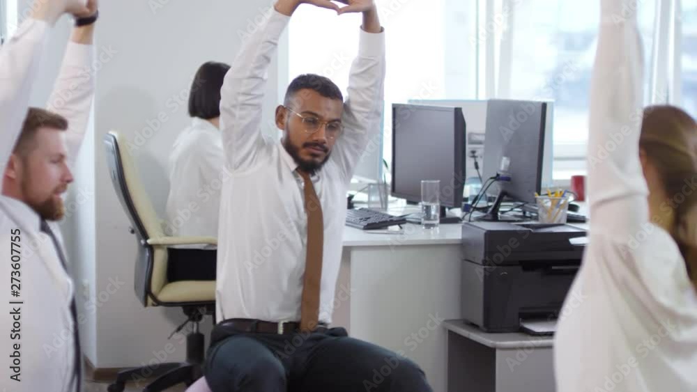 Tracking right of multiethnic colleagues sitting on fitballs in office and stretching their arms and shoulders