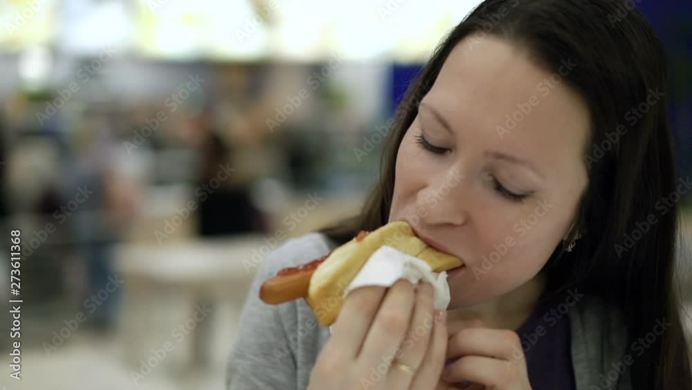 Young pretty woman eating tasty hot dog at fast food restaurant. Close-up hungry woman chewing at food court unhealthy food in shopping mall. Eating cheap snack, addicted to high calorie junk food