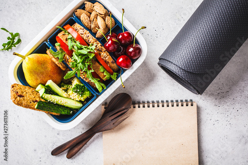 Sport food concept. Lunch box with heathy food, notebook and mat on a gray background.