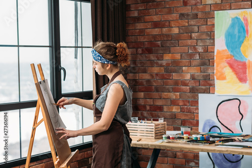 Home art studio. Side view of young female painter drawing on easel. Window, brick wall with abstract artworks in background. photo