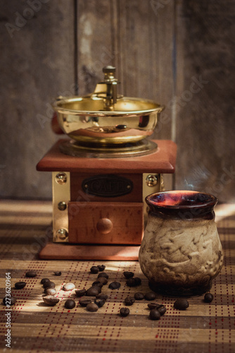 A Cup of black hot aromatic coffee with steam and an old vintage coffee grinder with a scattering of whole roasted coffee beans on a wooden table. Cozy rustic still life.