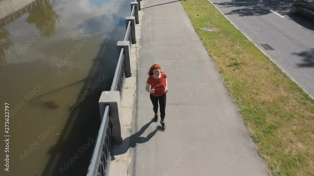 Shooting from the air red-haired sports woman enthusiastically running along the embankment of the city river Shooting from the air red-haired sports woman enthusiastically running along the embankment of the city river