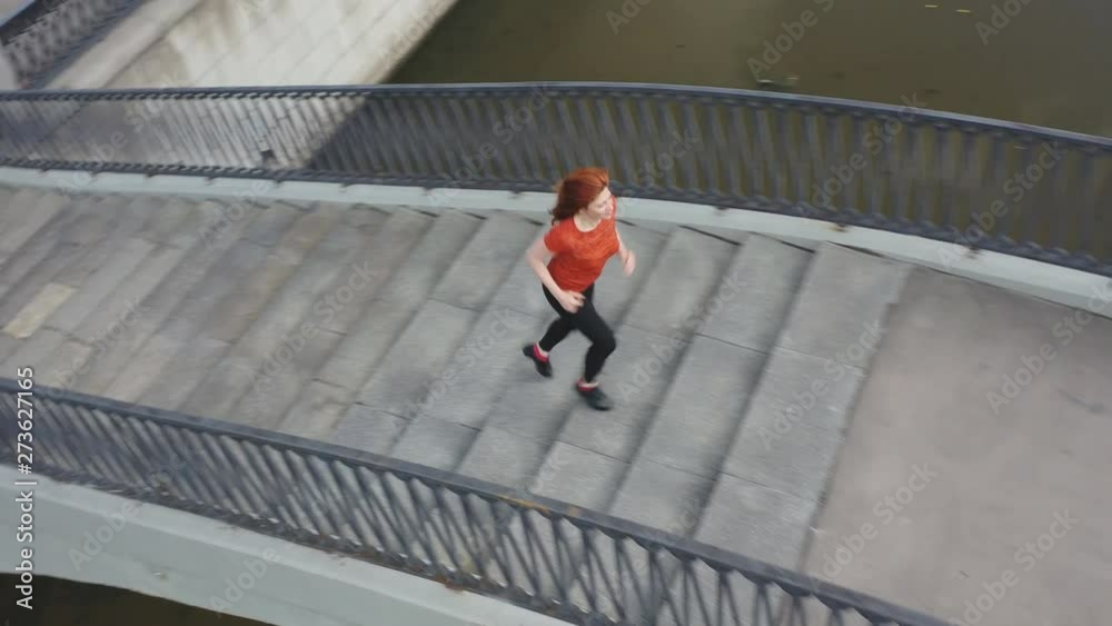 Shooting from the air red-haired sports woman enthusiastically running along the embankment of the city river Shooting from the air red-haired sports woman enthusiastically running along the embankment of the city river