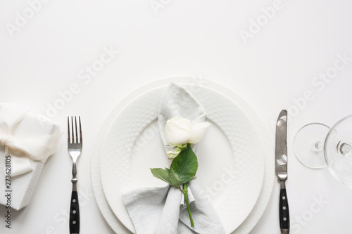 White table setting with bouquet rose, gift, dishware, silverware on holiday white table. Top view. Romantic and wedding anniversary. Space for text.
