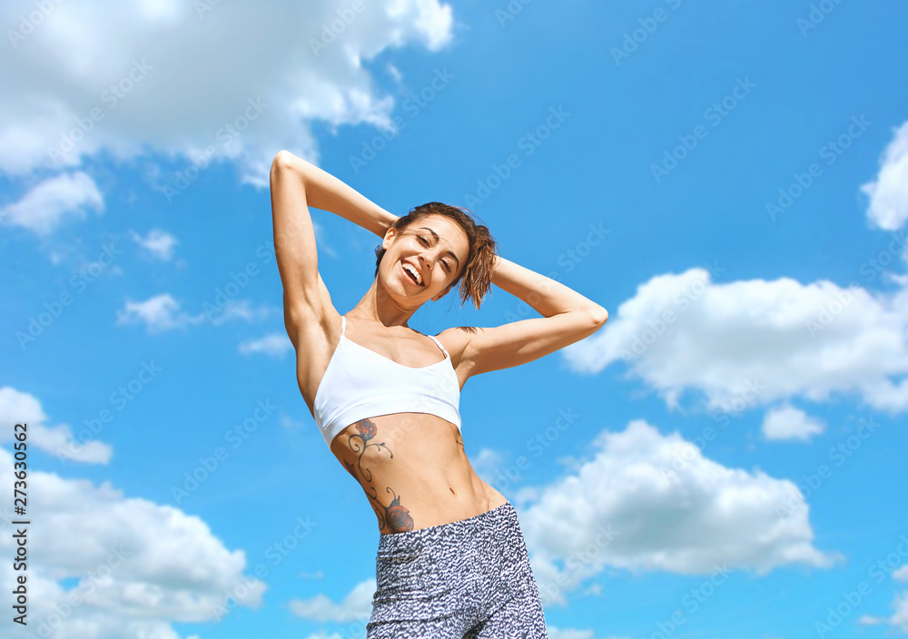 Happy sports woman with tanned fit body and tattoo posing in sport wear against blue sky with clouds. Healthy smiling female enjoying sunny summer day. Summertime concept. Happy sports woman with tanned fit body and tattoo posing in sport wear against blue sky with clouds. Healthy smiling female enjoying sunny summer day. Summertime concept.