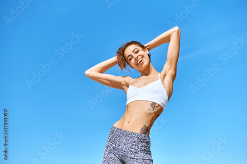 Happy sports woman with tanned fit body and tattoo posing in sport wear against blue sky. Healthy smiling female enjoying sunny summer day. Summertime concept. © vitaliymateha