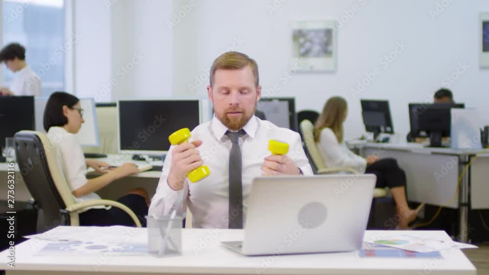Medium shot of Caucasian businessman with red hair sitting at desk at laptop, typing then taking dumbbells and starting doing exercises while his colleagues working on background Medium shot of Caucasian businessman with red hair sitting at desk at laptop, typing then taking dumbbells and starting doing exercises while his colleagues working on background