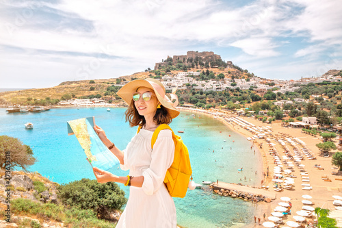 Happy asian woman with map and backpack wearing big vacation hat at the background of paradise view to Lindos town and beach on a Rhodes Island, Greece