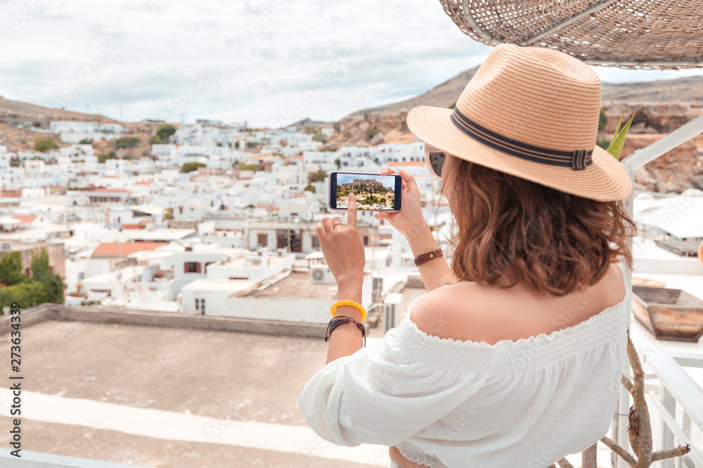 Young elegant woman tourist taking photos of ancient white town with her smartphone