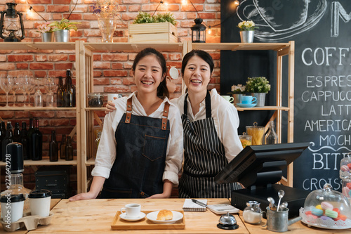 Small startup business owner concept. two successful young baristas women standing in bar counter in cafe. happy coffeehouse waitresses in aprons smiling confidently to camera in coffee shop. photo
