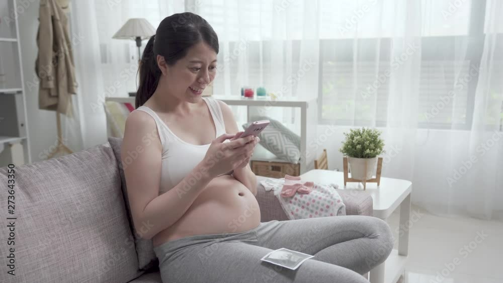 beautiful pregnant woman wearing sport bra sitting on couch using cellphone. young mother joyful texting message and chatting online with husband. bare tummy lady holding sonogram taking picture. beautiful pregnant woman wearing sport bra sitting on couch using cellphone. young mother joyful texting message and chatting online with husband. bare tummy lady holding sonogram taking picture.