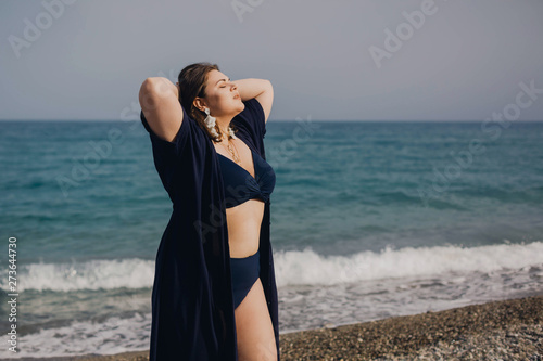Attractive busty curvy woman in a blue swimsuit resting on the beach. Stylish accessories, fringe, fashion for plus size, beautuful sea. Bodypositive, natural authentic beauty, resort, summer vacation