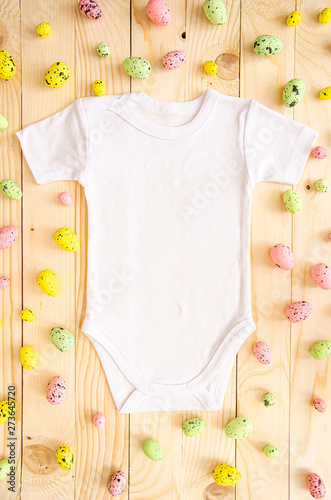Top view white baby romper bodysuit on wooden background with easter eggs. Copy space for lettering or your text. Flat lay mockup photo