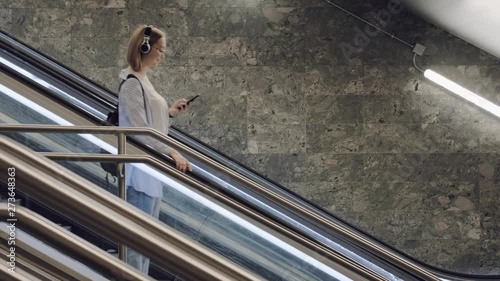 Young beautiful girl goes down the escalator in the subway and looks into the smartphone. A student with headphones goes to the university. Listening to music on headphones in the smartphone.