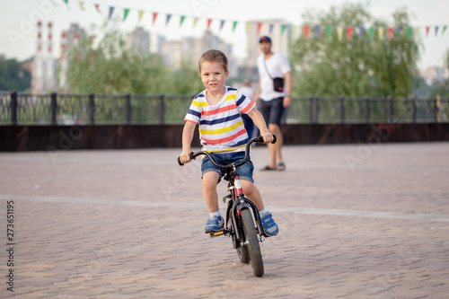Fototapeta Naklejka Na Ścianę i Meble -  Active preschool kid boy without helmet biking on bicycle in the city. Happy child in colorful clothes and city traffic. Safety and protection for children. Active leisure for kids outdoors