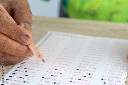 Close-up image of elderly asian woman hands holding pencil for filling in standardized test with drawing selected choice on answer sheets on wood table. Education and Lifelong learning concept.