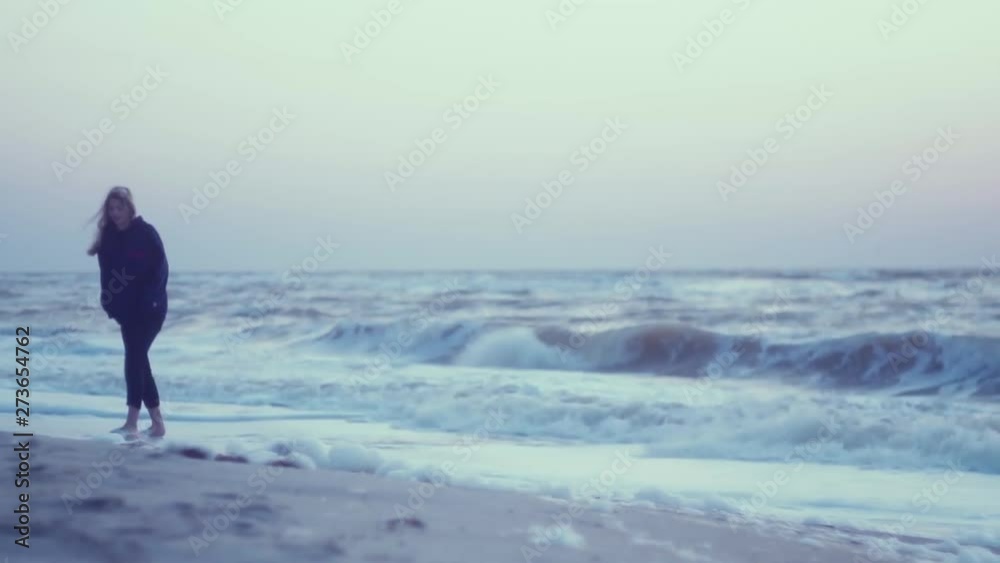 Beautiful girl walks at dawn on the surf, on the wet sand, on the shore by the sea. Pink sky, waves, wind in the background Beautiful girl walks at dawn on the surf, on the wet sand, on the shore by the sea. Pink sky, waves, wind in the background