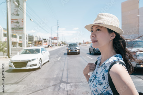 Young asian woman with backpack crossing road at pedestrian crosswalk. smiling girl tourist in straw hat walking in sunny las vegas boulevard city street in hot summer. blurred cars on background.