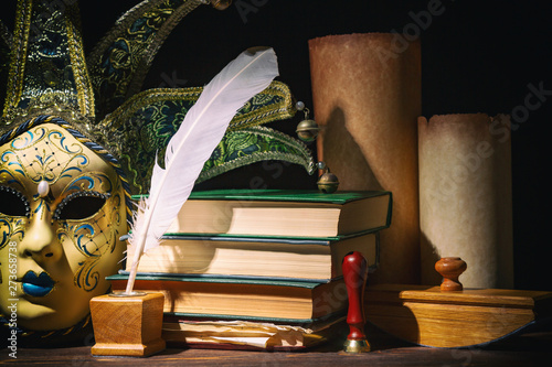 Theater, literature and drama concept. Venetian mask with old inkwell, feather, quill, scrolls, books and seal on wooden table. Vintage still life.