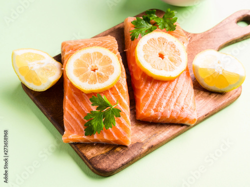 Salmon fillet on wooden board with lemon slices, parsley and pink salt. Pastel green background. Healthy fish meal to be cooked. Protein source.