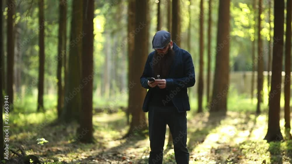 Vape bearded man. An adult white short-haired man with a very long beard in a cap and glasses smokes an electronic cigarette and talks to smartphone outside in the forest on a sunny day. Bad habit. Vape bearded man. An adult white short-haired man with a very long beard in a cap and glasses smokes an electronic cigarette and talks to smartphone outside in the forest on a sunny day. Bad habit.
