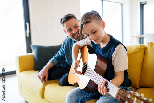 Mature father with small son sitting on sofa indoors, playing guitar.