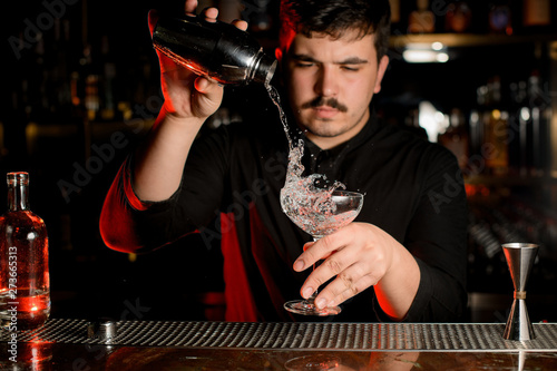 Professional bartender pouring a transparent alcoholic drink into the cocktail glass from the steel shaker